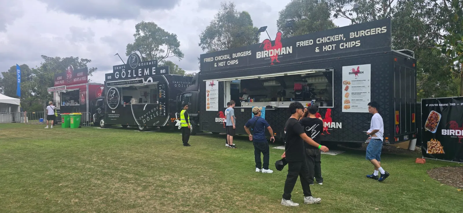 A line of food vendors serving at a festival, with a staff member in hi-vis managing the precinct